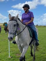 Person riding a gray horse in a grassy field, wearing a helmet and purple shirt.