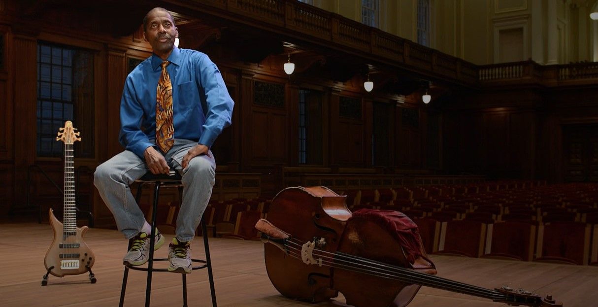 Avery Sharpe sitting on a stool in front of a double bass and a guitar .