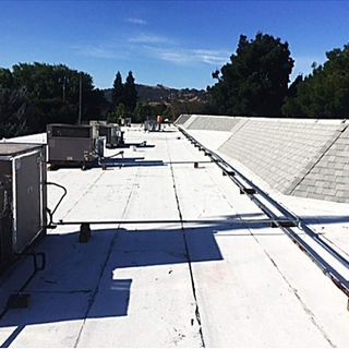 Rooftop with HVAC units and metal conduit. Bright white surface, trees, and blue sky in background.