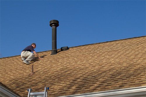Man on a roof near a black chimney, checking something. Brown shingles, blue sky, and ladder visible.