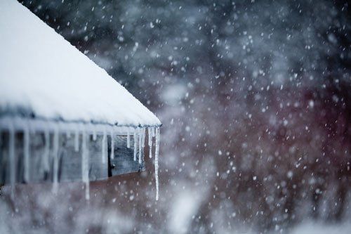 Snow-covered roof with icicles; heavy snowfall in the blurry background.
