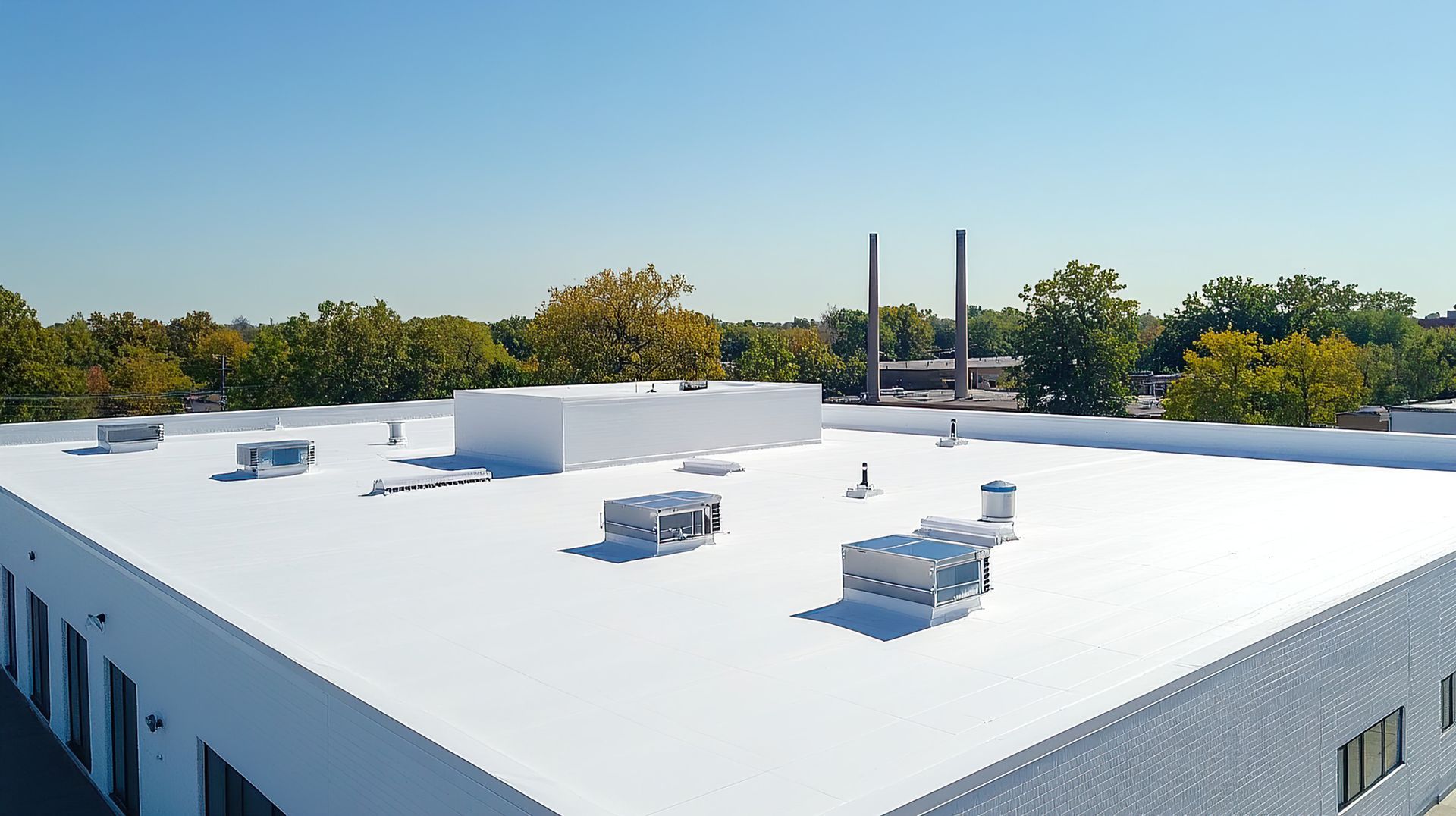 White commercial flat roof with vents, against blue sky and tree line.