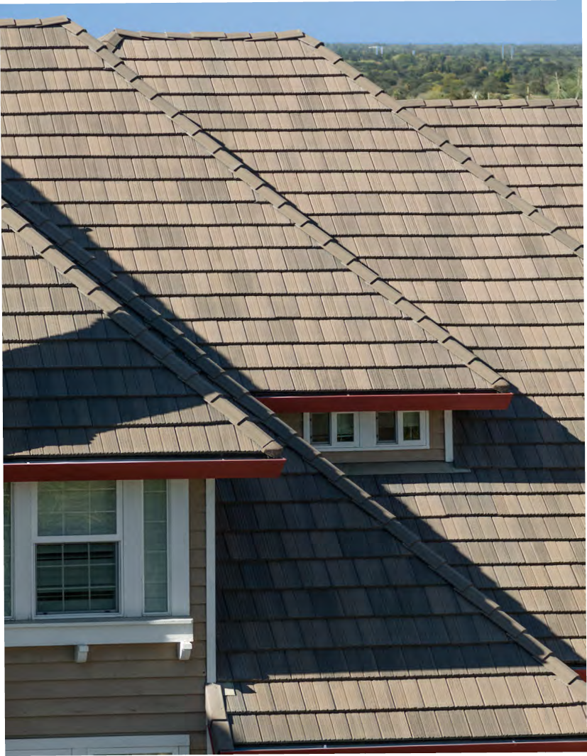 A close-up view of a house roof featuring textured brown tiles, eaves, and windows under a clear sky.