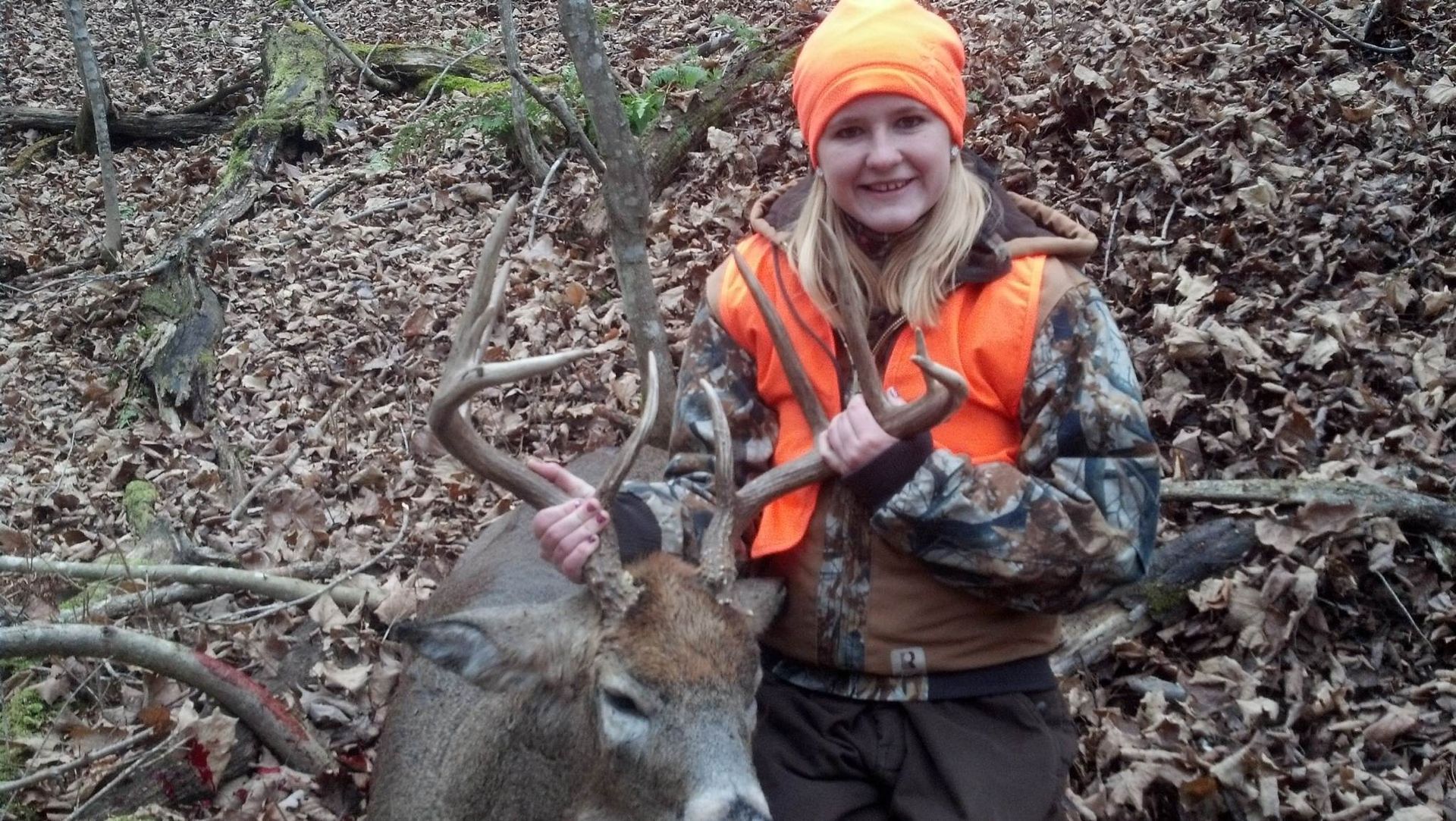 Woman in orange vest and hat holds antlers of a harvested deer in a wooded area.