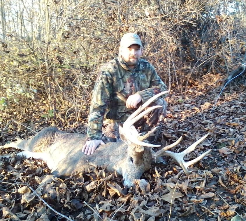 Man in camouflage kneels by a harvested buck with large antlers on a bed of leaves.