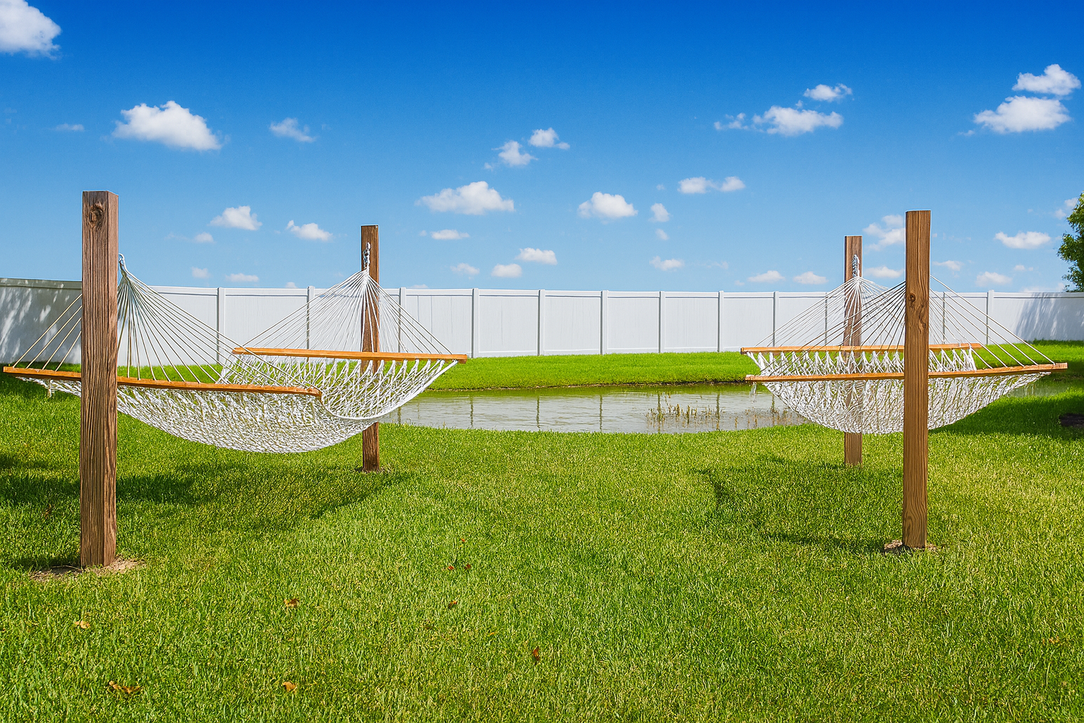 Two white hammocks suspended between wooden posts on a green lawn with a blue sky background.