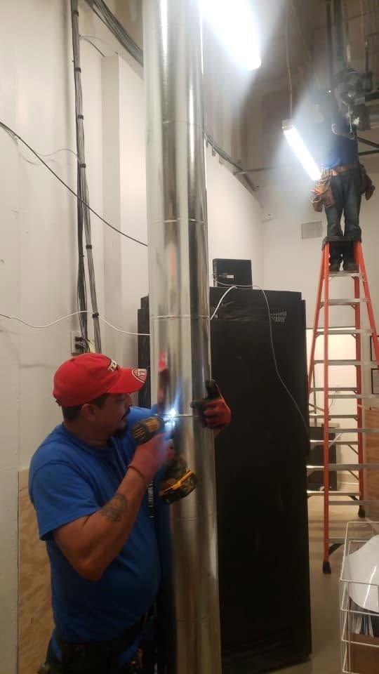 Workers on ladders installing equipment in a narrow utility corridor, with one drilling near a wall.