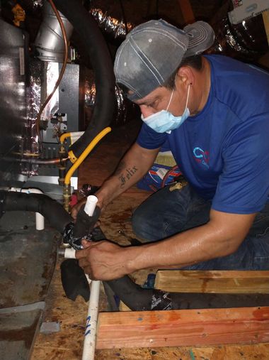 Technician in blue shirt and mask working on exposed pipes in a cramped utility space
