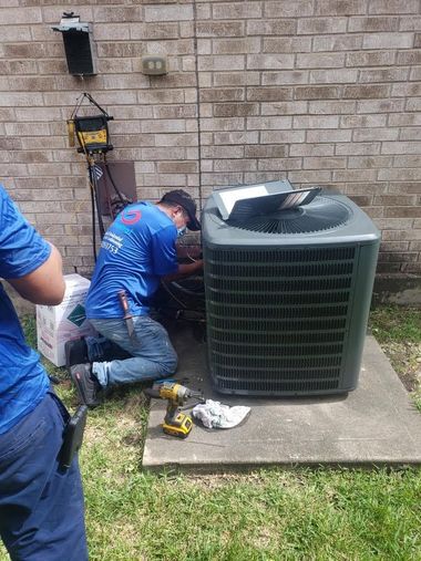 Technician kneeling beside outdoor air conditioner unit, working with tools against a brick wall.