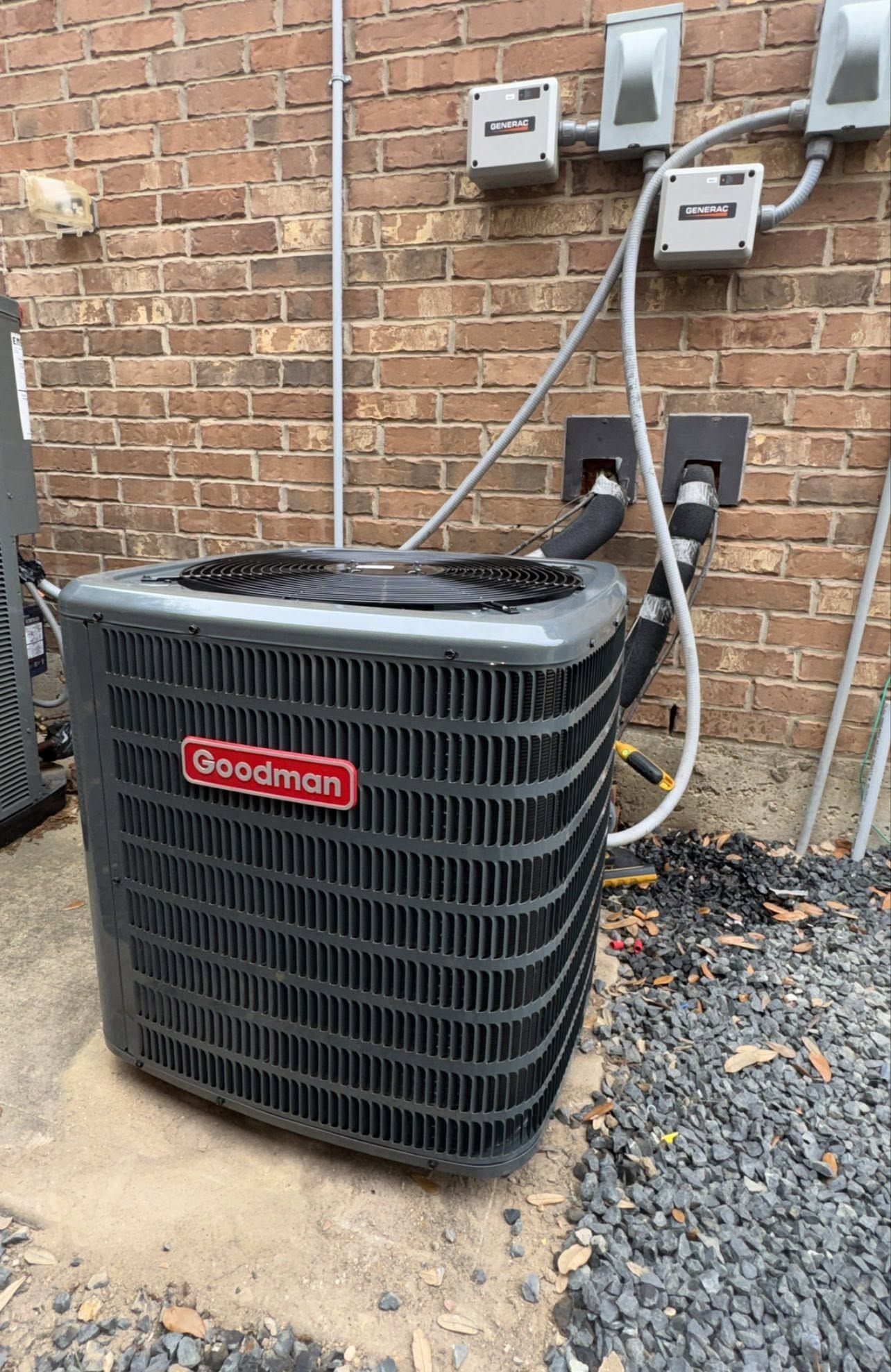 Black Goodman outdoor air conditioner unit beside a brick wall with utility meters and pipes