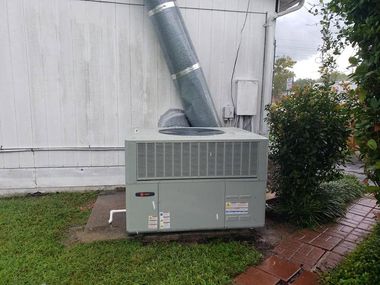 Outdoor gray HVAC unit with metal duct beside a white building and brick walkway