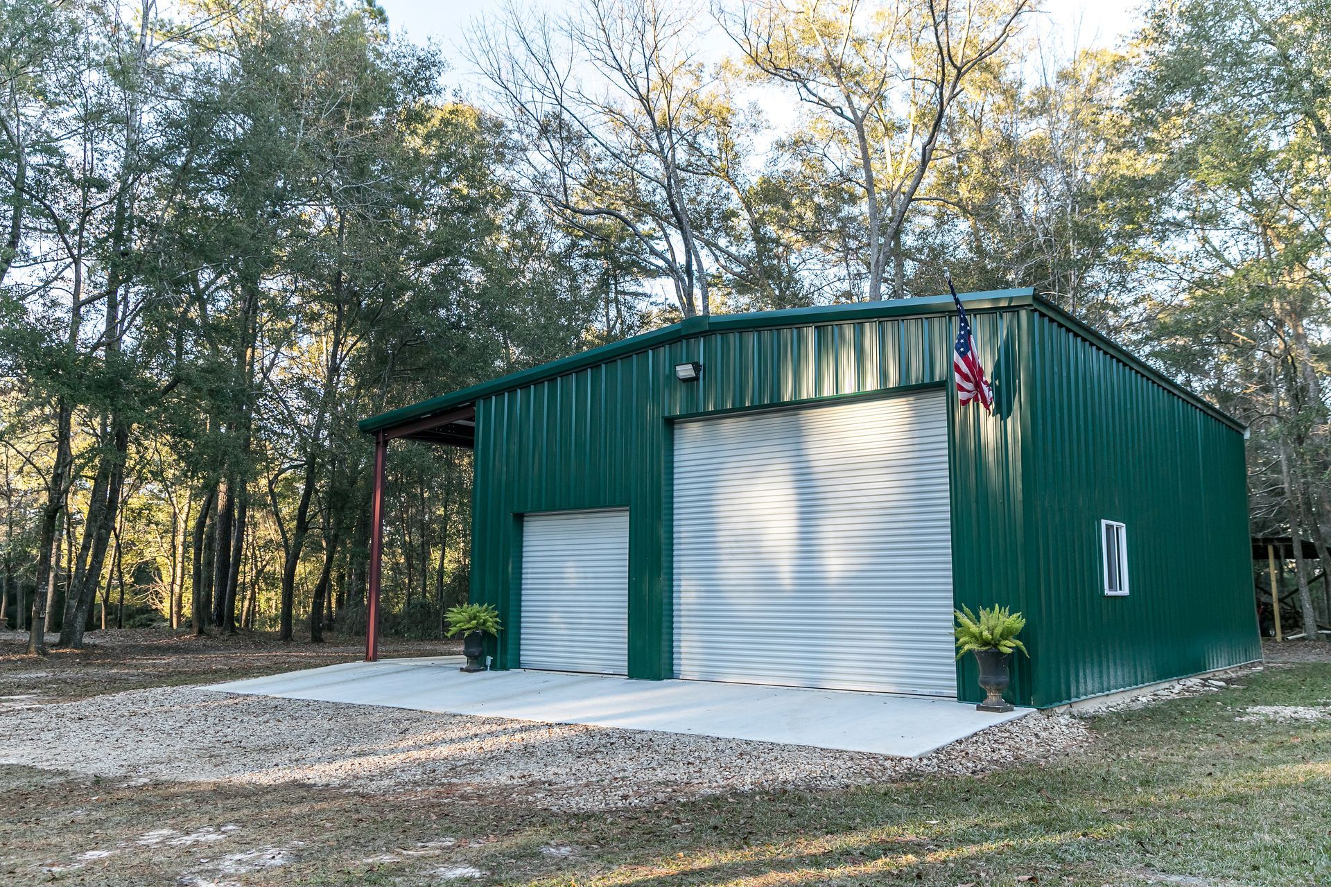 A green metal building with a white garage door is in the middle of a forest.