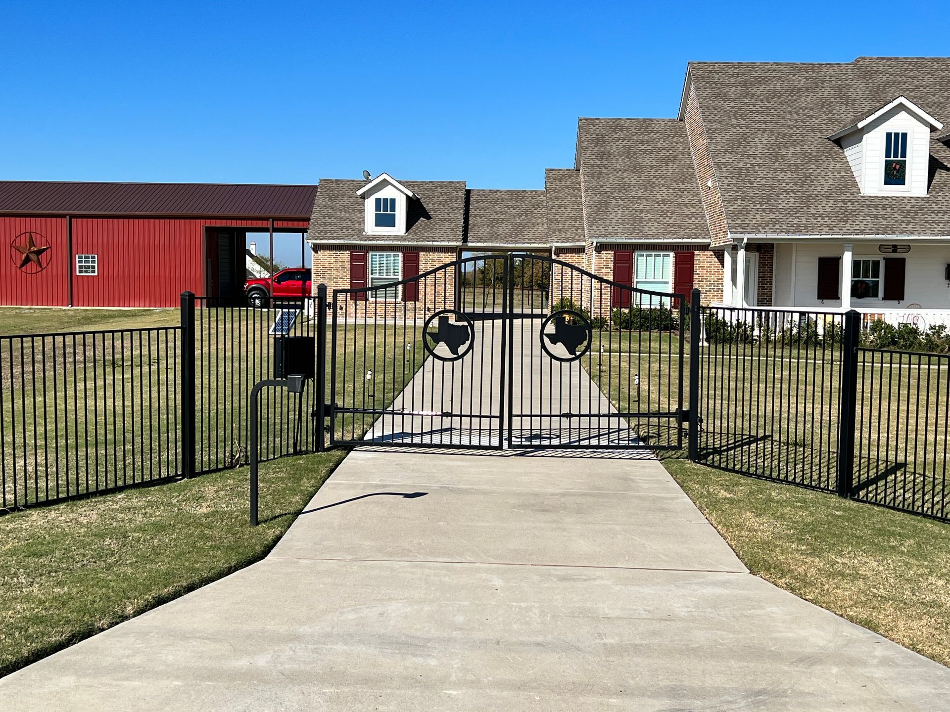A fence surrounds a driveway leading to a house with a red barn in the background