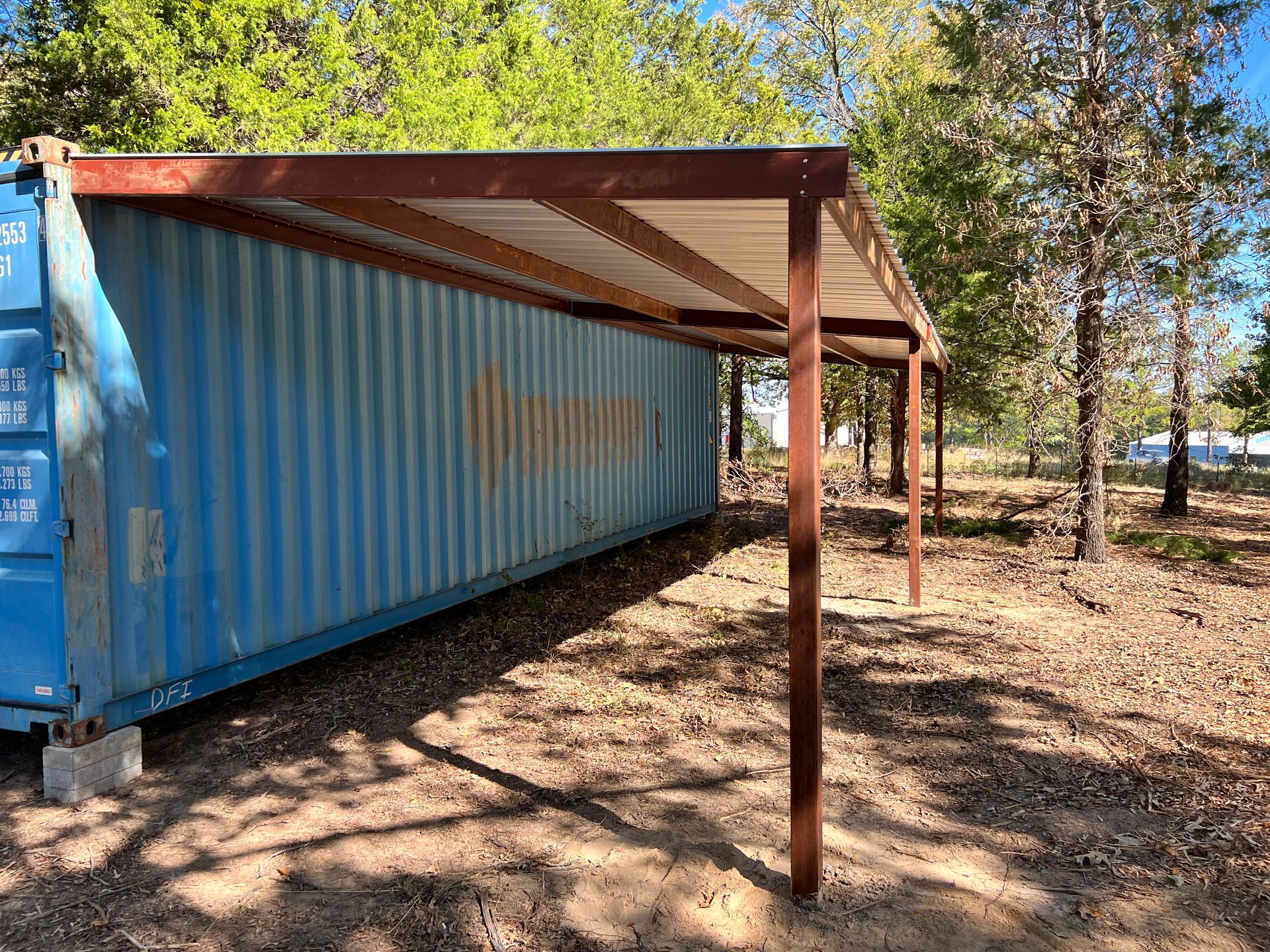 A blue shipping container is sitting under a wooden structure in a field.