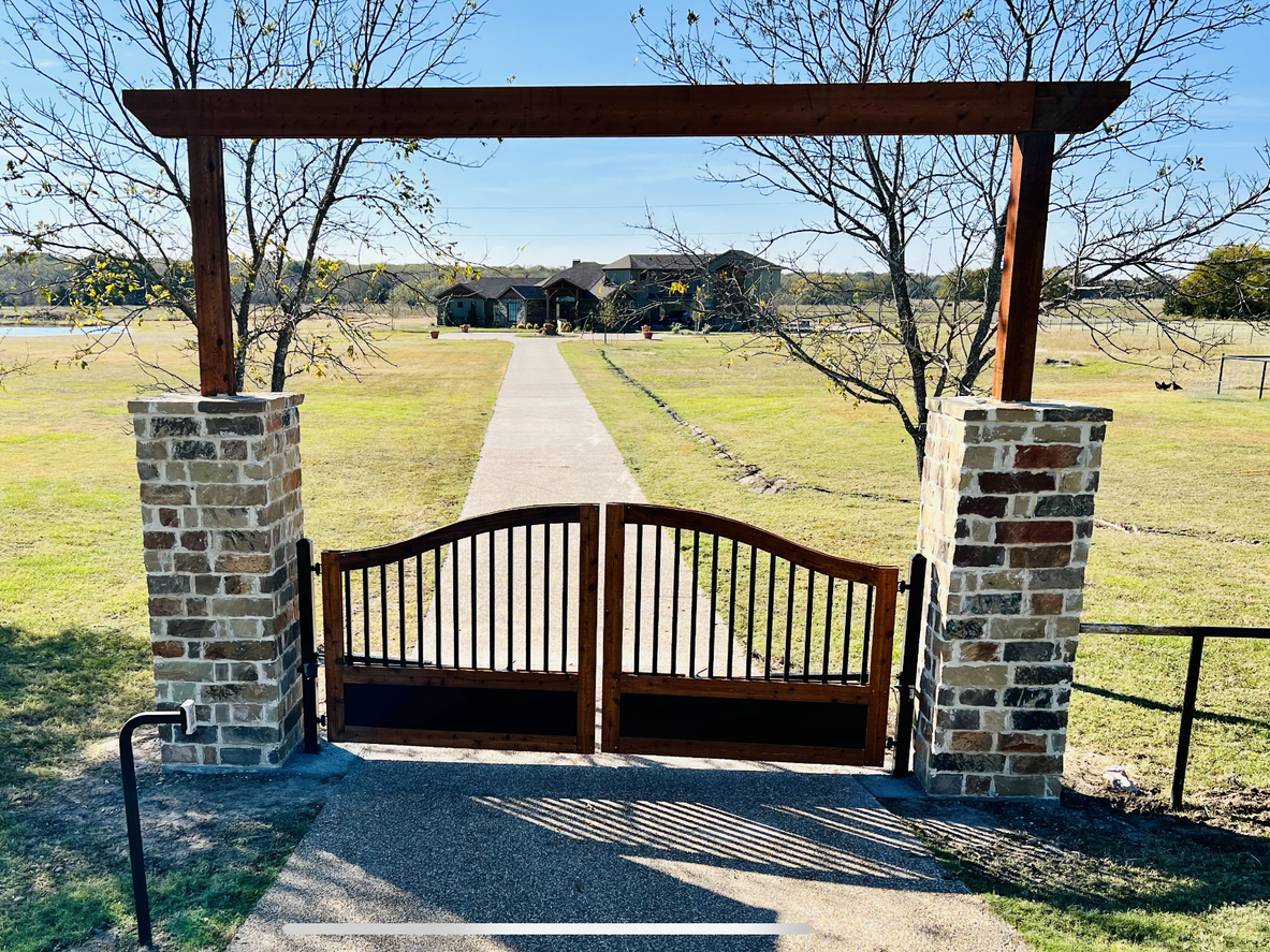 A wooden gate is surrounded by brick pillars and a stone walkway leading to a house.