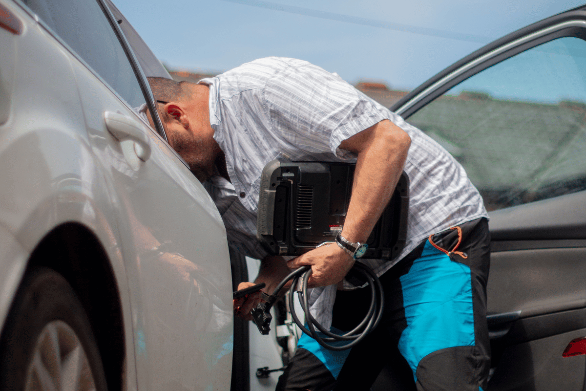 Man peering into a car with a black device, possibly for diagnostic purposes, in daylight.