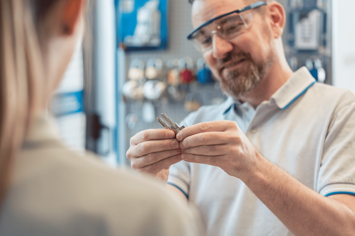 Man in safety glasses inspecting a key, smiling at a customer in a shop.