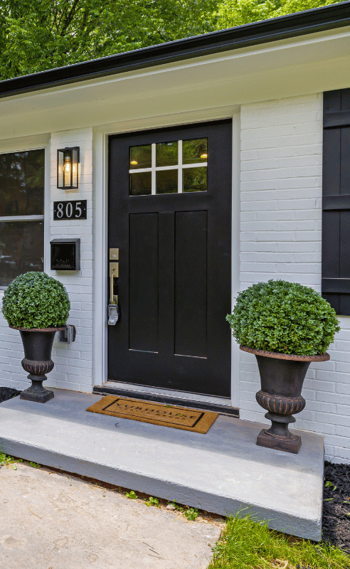 Black door with white trim, brick facade, and potted plants.