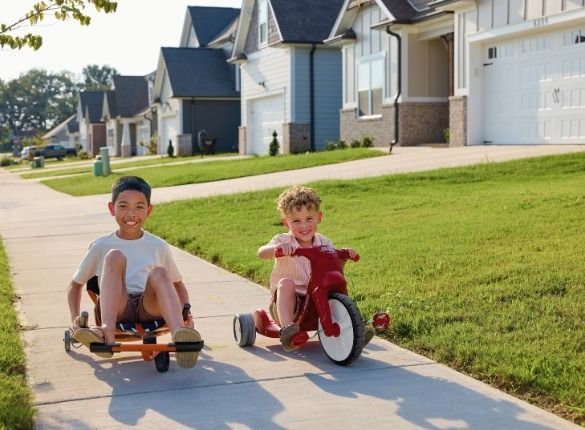 Two children on a sidewalk, one on a scooter, the other on a red tricycle, in front of houses.