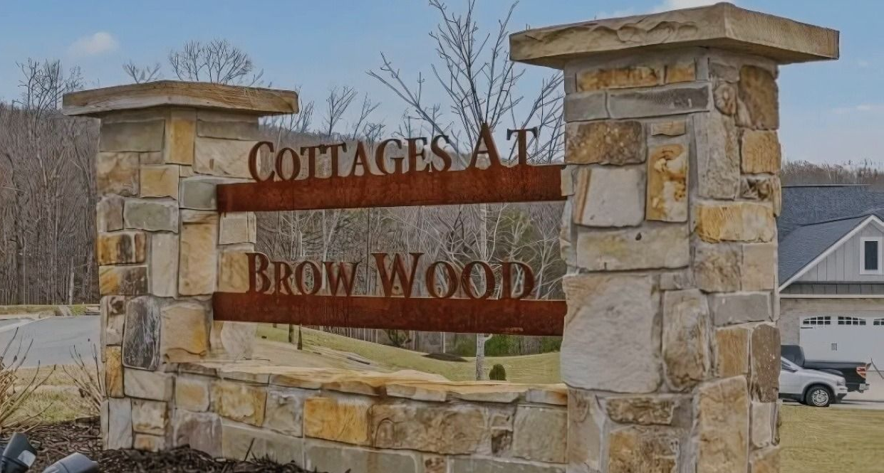 Sign for "Cottages at Brow Wood" in front of a stone structure, brown text on a rust-colored panel.