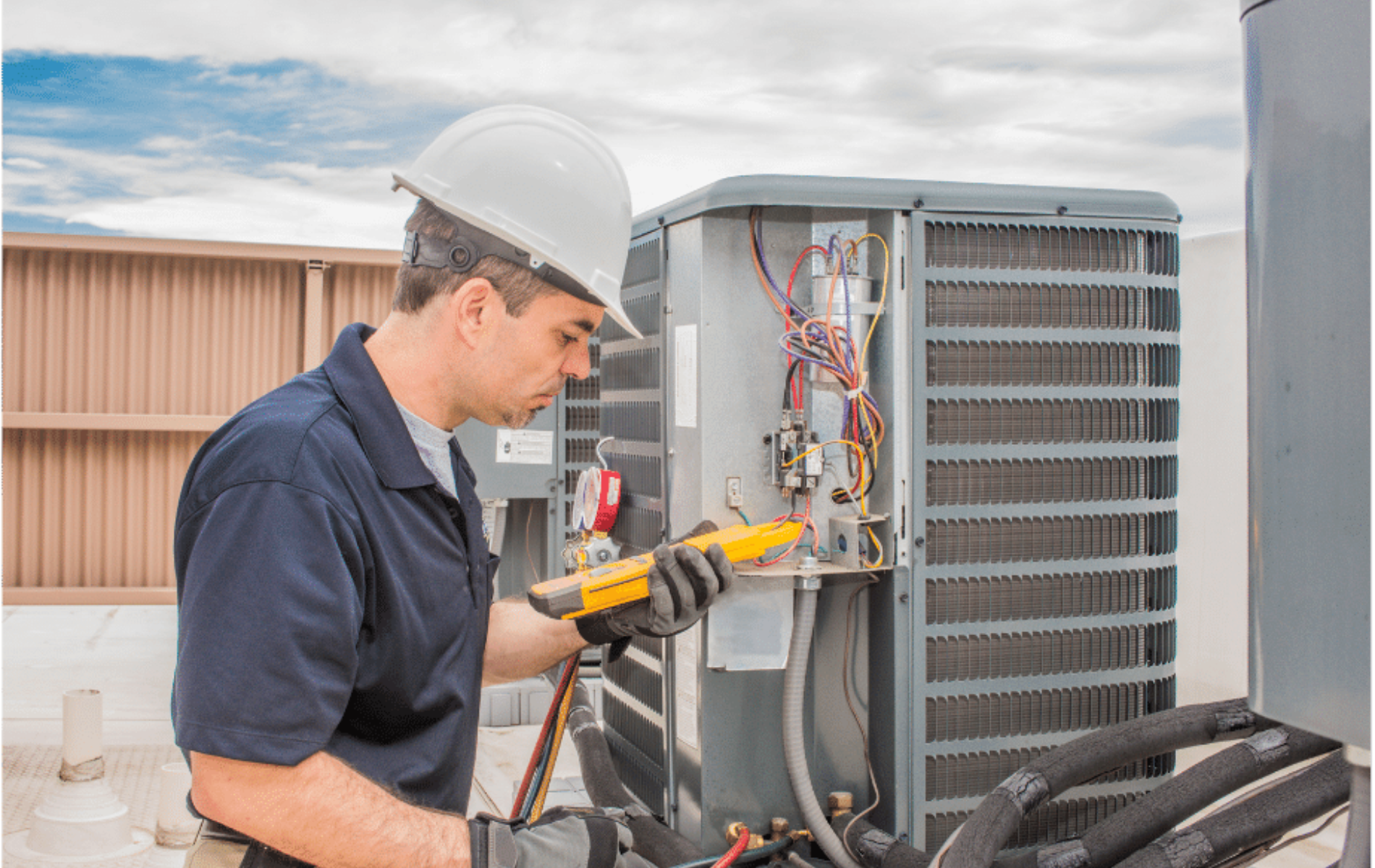 HVAC technician in a hard hat inspects air conditioning unit with a multimeter on a rooftop, sunny day.