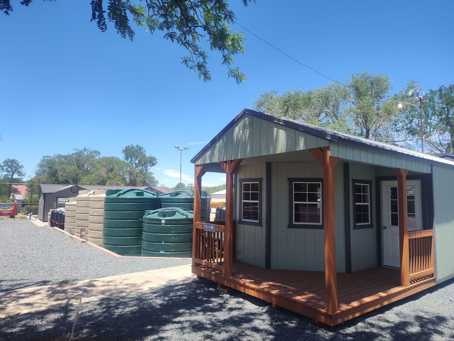 Small green cabin with wooden porch beside stacked water tanks on a gravel lot under a clear blue sky