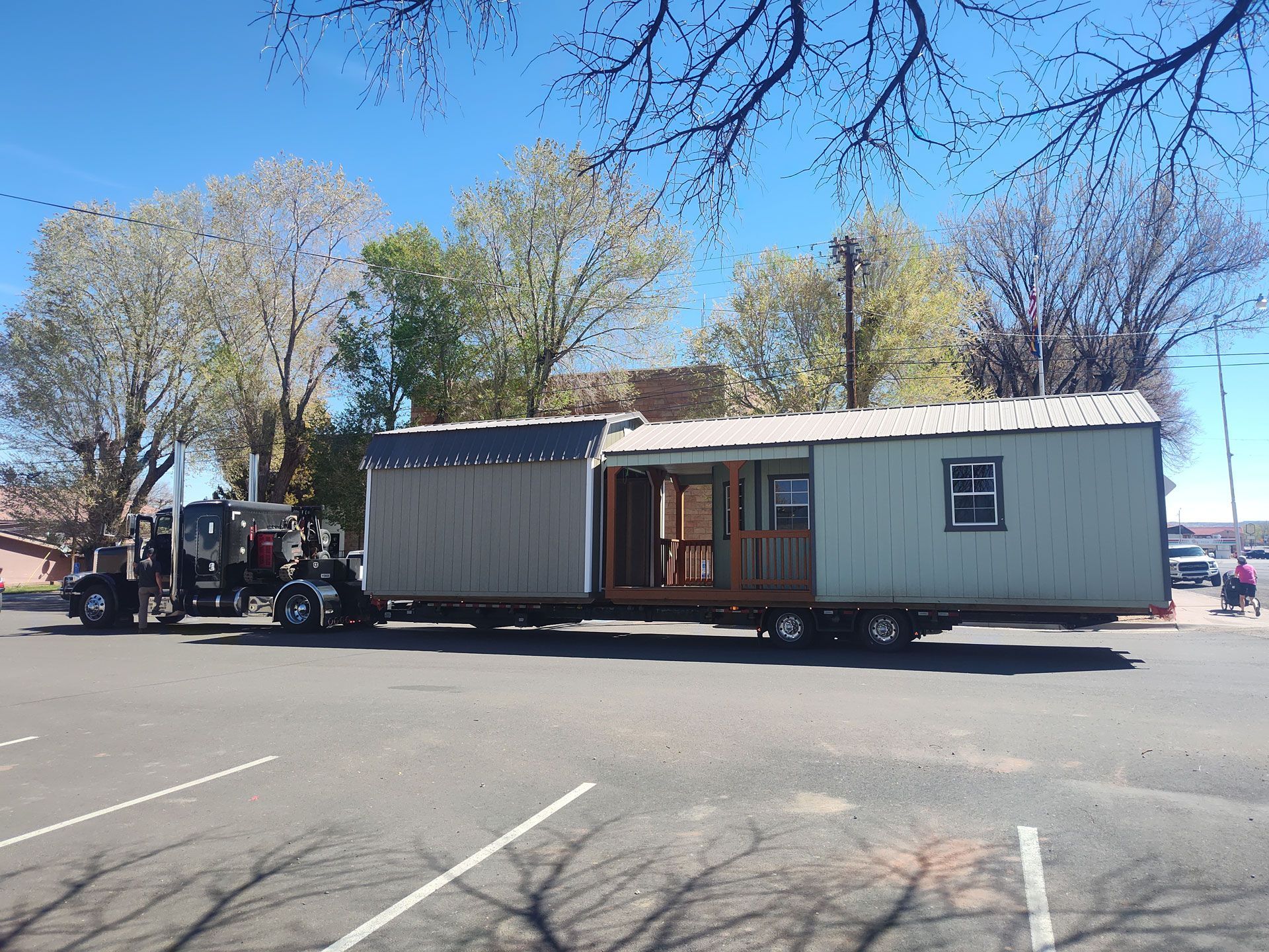 Large mobile home being transported on a truck in a parking lot under a clear blue sky.
