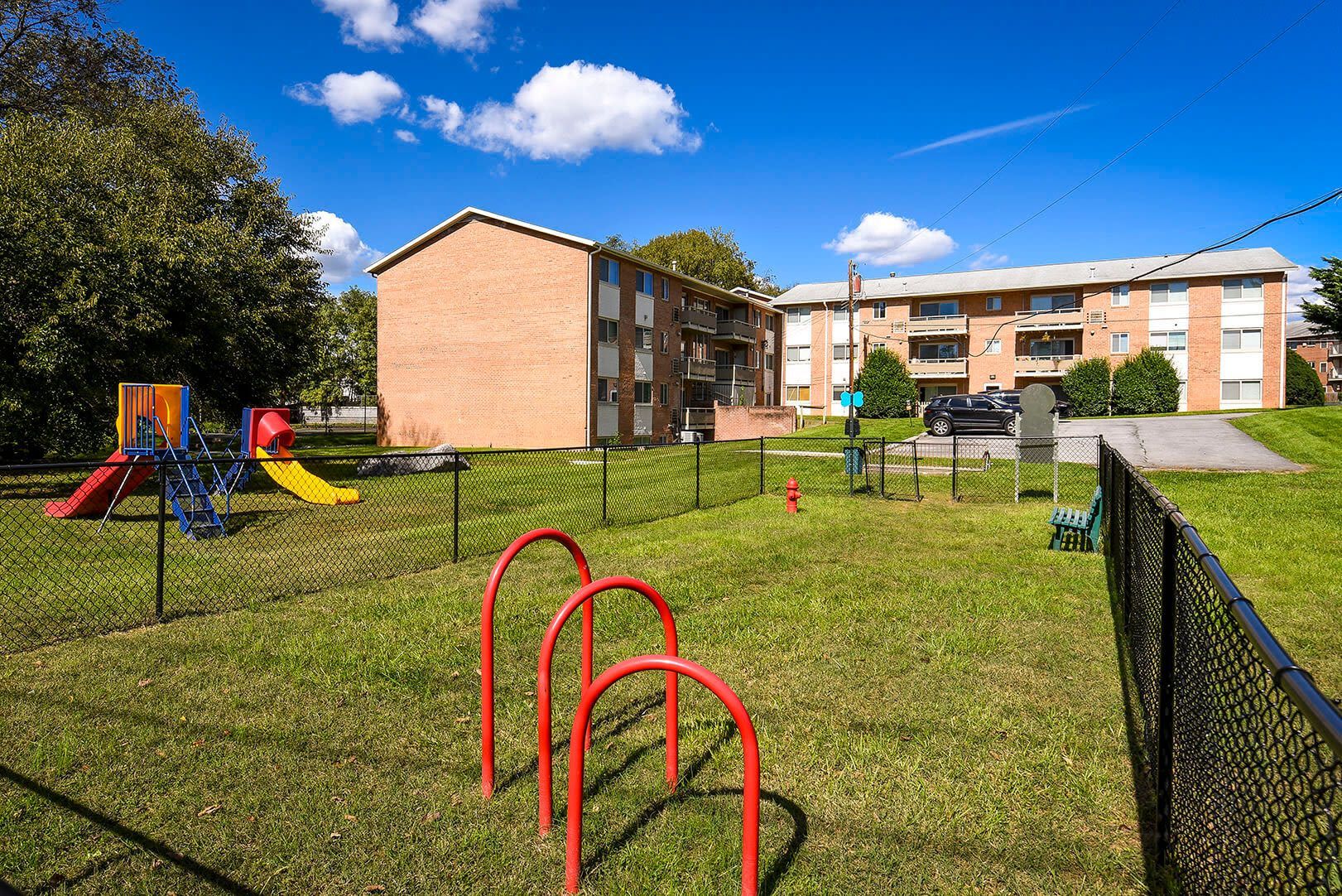 Fenced playground with red metal arches in foreground and brick apartment buildings behind at The Ridge Apartments in Hagerstown, MD
