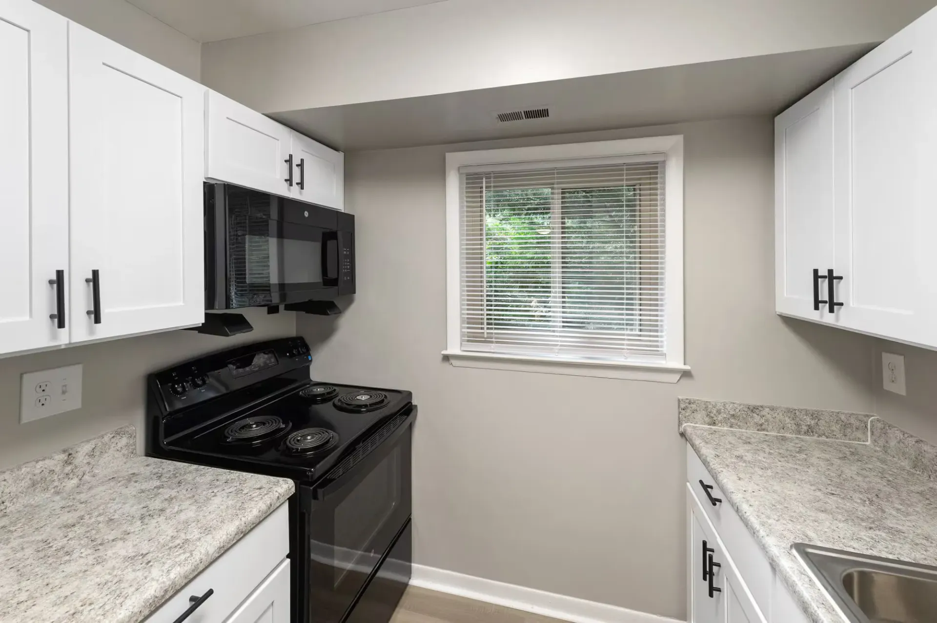 Compact apartment kitchen with white cabinets, black stove and microwave, speckled countertops, and a window with blinds at The Ridge Apartments in Hagerstown, MD