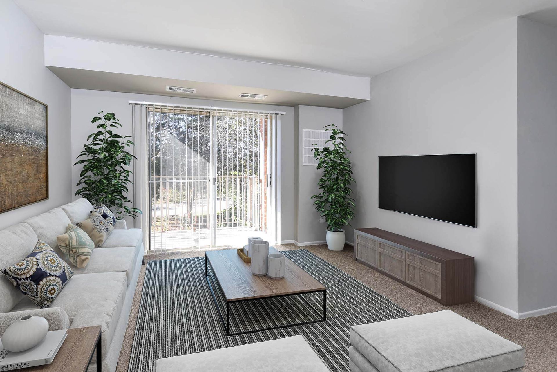 Living room in an apartment with a white sofa, coffee table, TV, sliding glass door, and tall potted plants at The Ridge Apartments in Hagerstown, MD