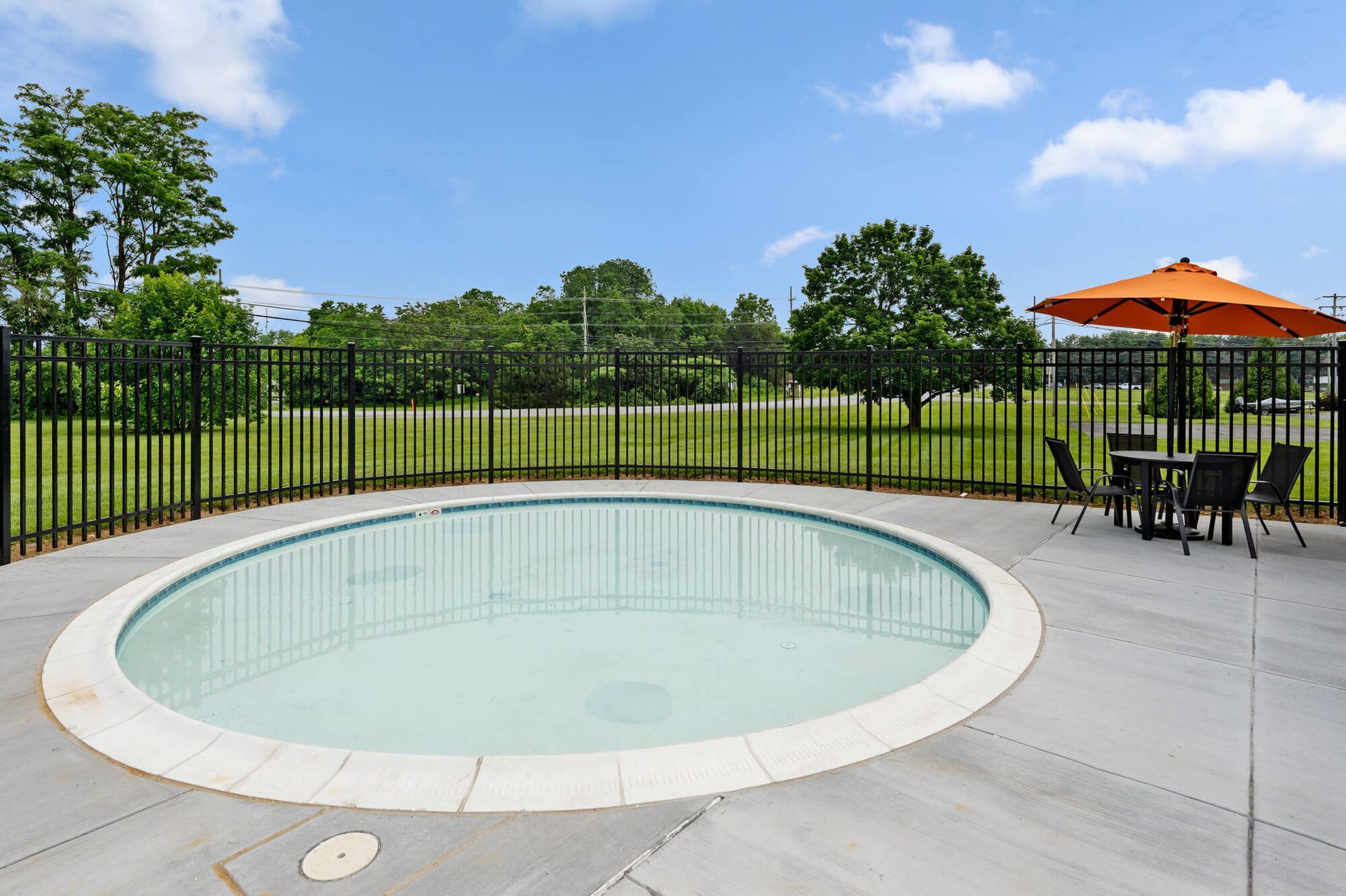 Circular outdoor pool with a black fence, concrete deck, and a table with an orange umbrella at The Ridge Apartments in Hagerstown, MD