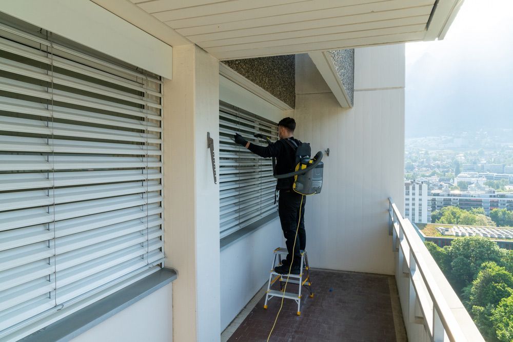 A Man is Standing on a Ladder Cleaning a Window With Blinds — Dazzling Windows & Pressure Cleaning in Tugun, QLD