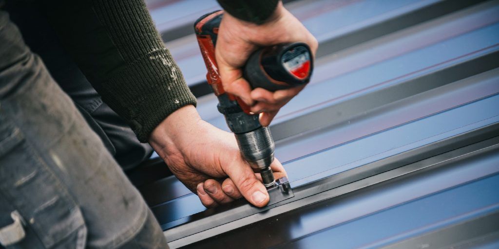 Hands using a drill to fasten a screw in a blue corrugated metal roof panel