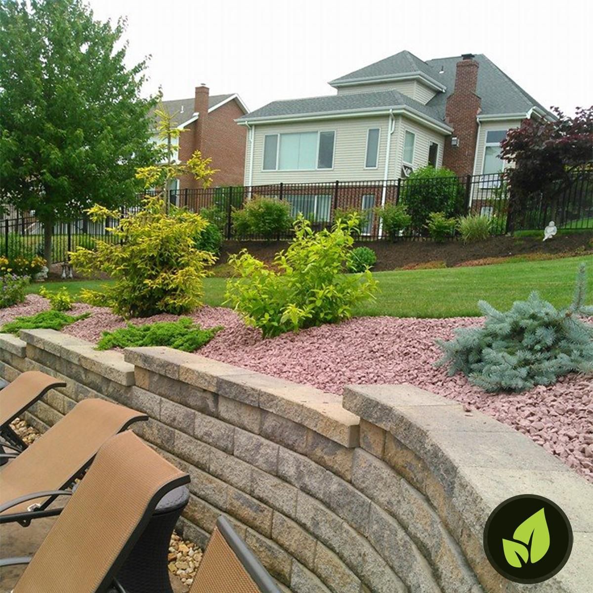 Curved retaining wall with landscaped beds of red rock, bushes, and a house in the background.