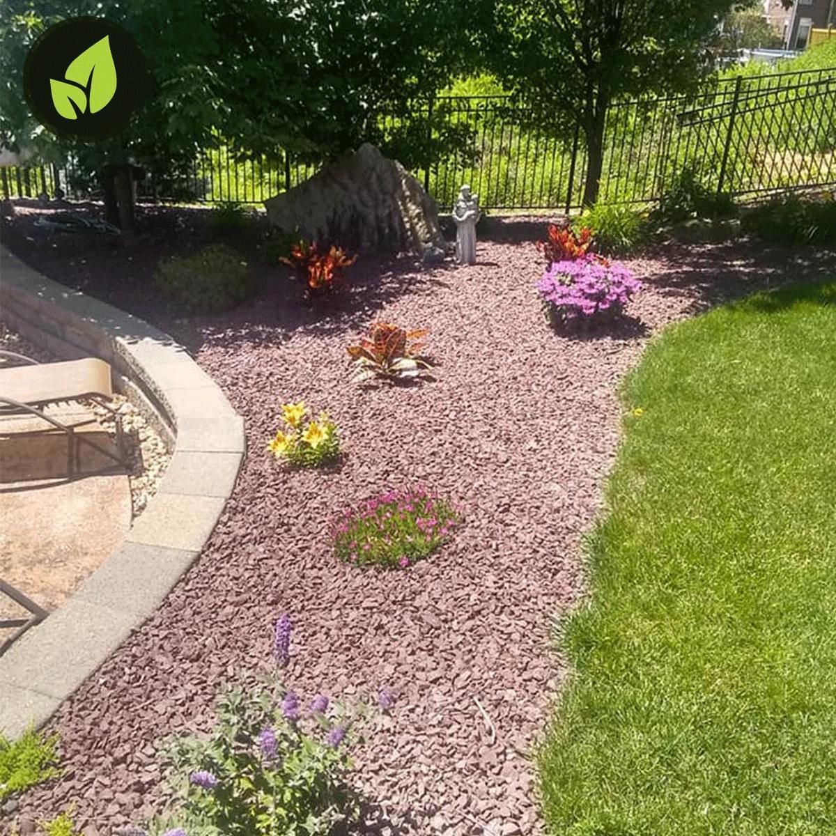 Landscaped garden bed with red mulch and colorful flowers, next to green lawn and stone patio.
