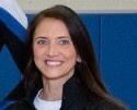 A woman in a black jacket is smiling in front of a blue wall.