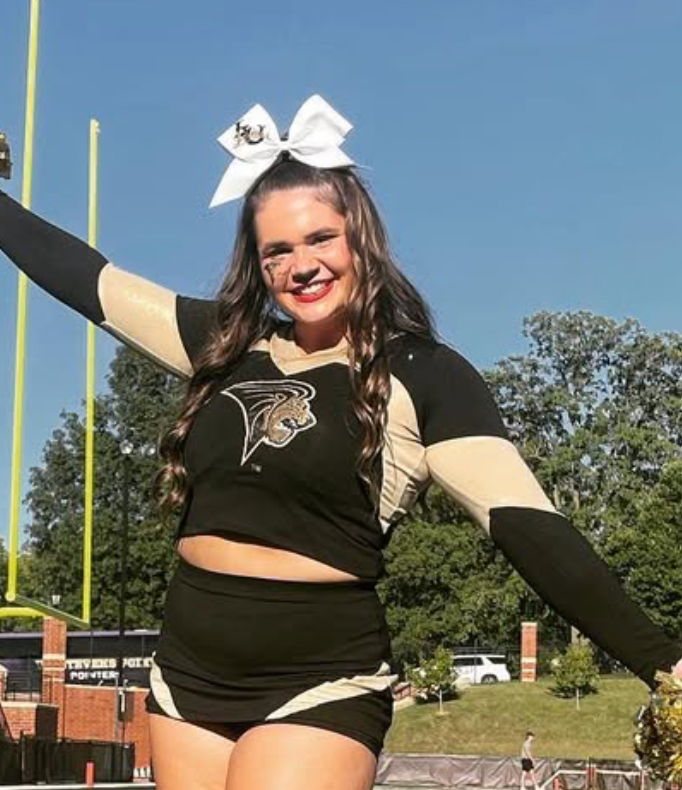 A cheerleader with a bow in her hair is standing on a field with her arms outstretched.