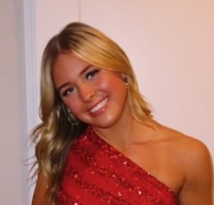 A woman is wearing a red one shoulder dress and smiling.