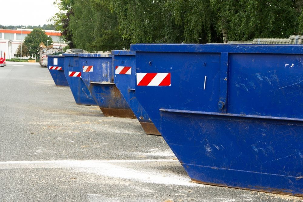 A row of blue dumpsters are lined up in a parking lot.