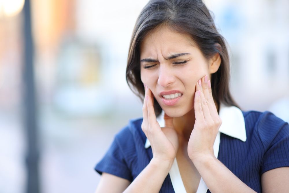 Woman with pained expression, hands on cheeks, in an outdoor setting, indicating possible toothache or jaw pain.