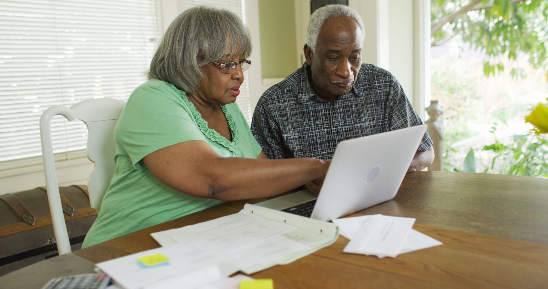 Two people sit at a table reviewing a laptop and papers in a bright room.