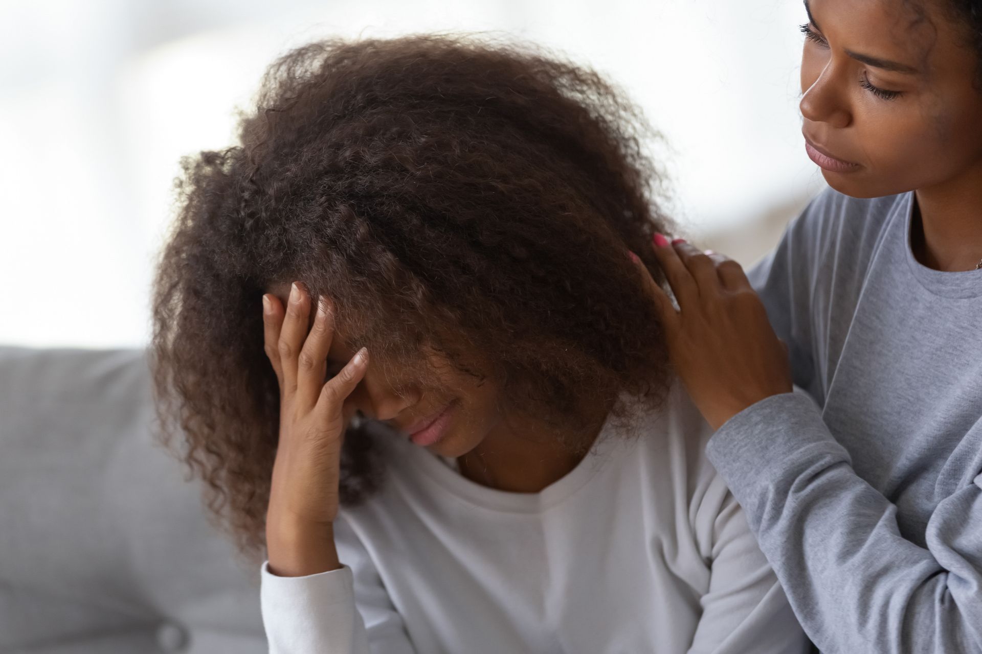 One person comforts another with a hand on their shoulder as they sit upset on a couch.