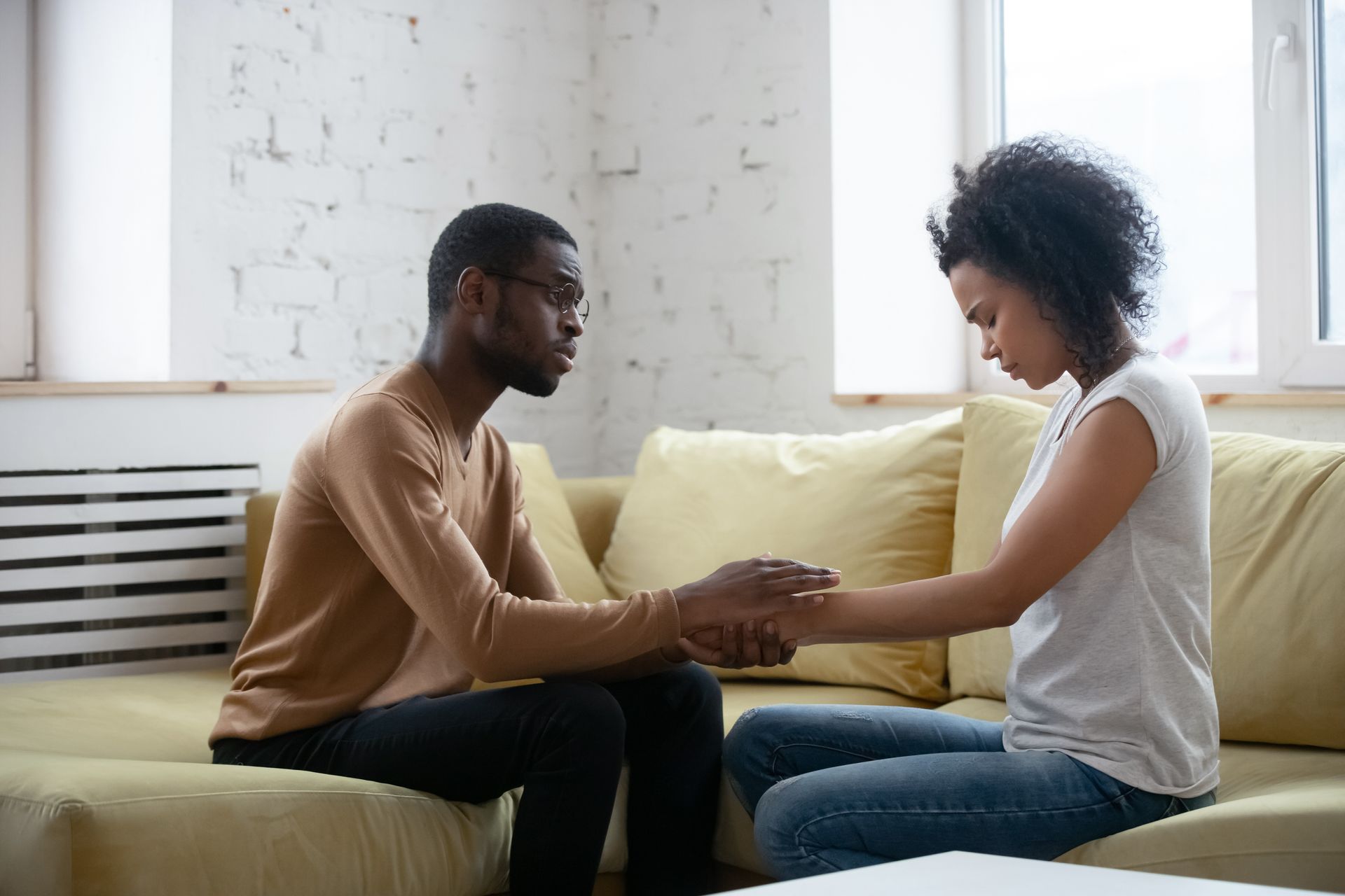 Two people sit on a couch, holding hands and facing each other in a bright room.