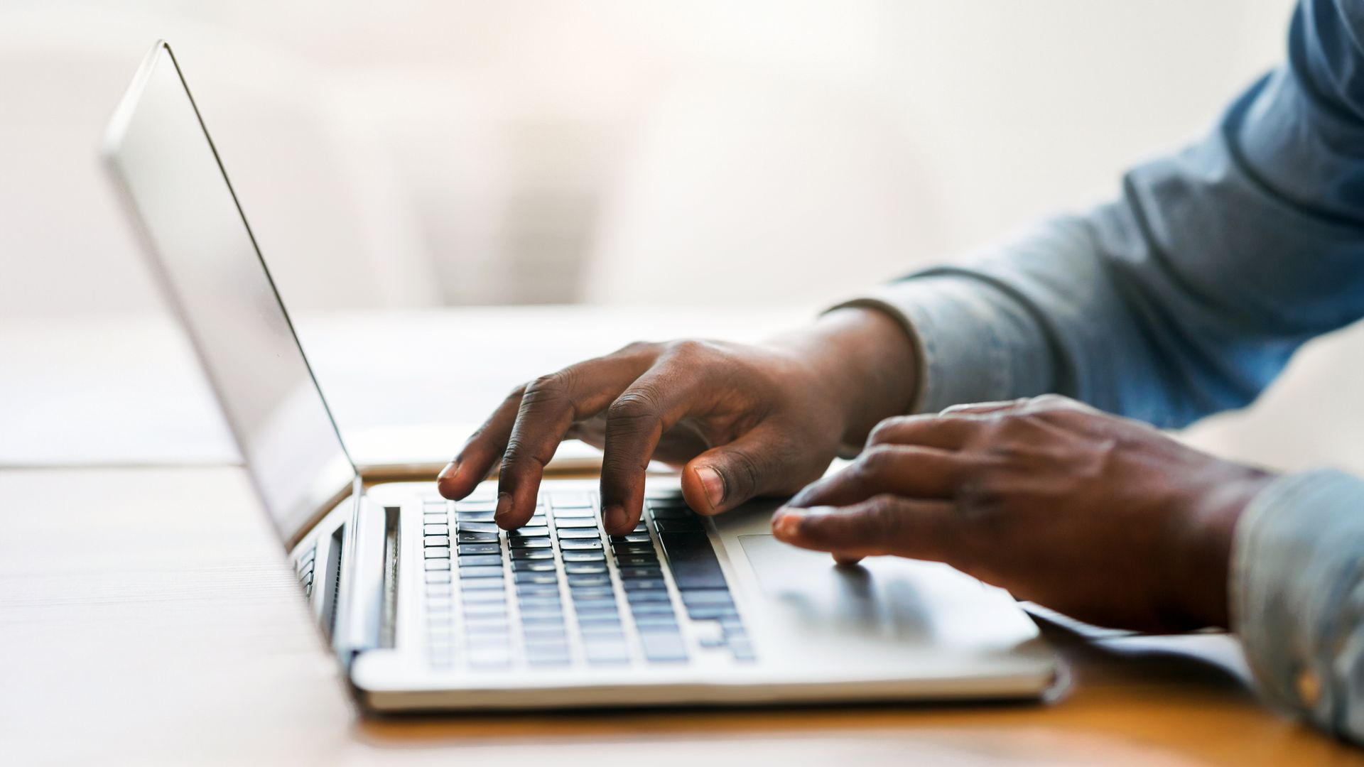 Hands typing on a silver laptop keyboard at a desk, with a bright blurred background.