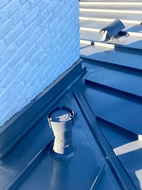 Close-up of a dark metal roof with a chimney and a small vent; white brick is visible.