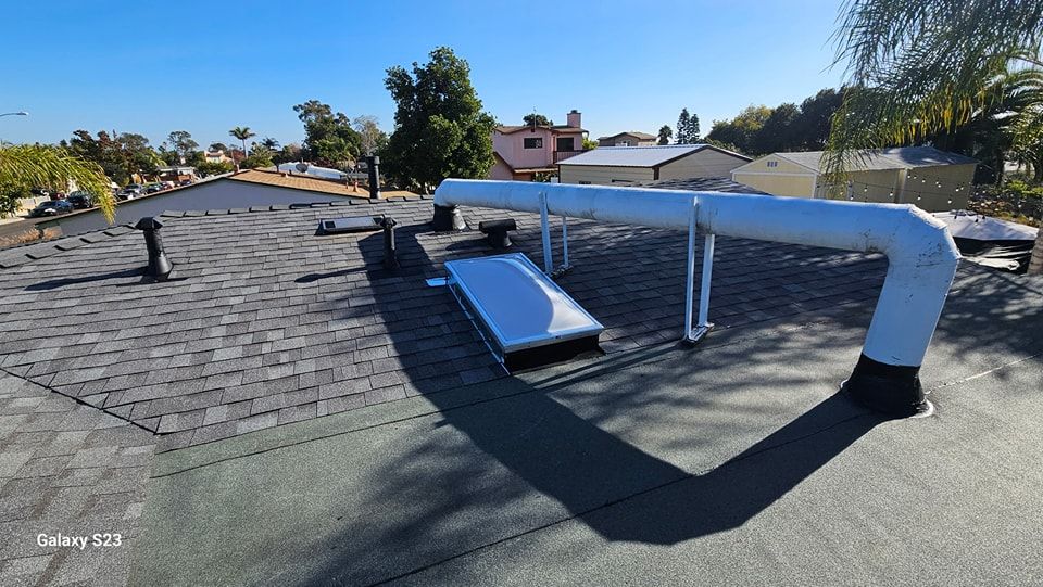 Rooftop view with dark shingles, vents, a skylight, and white pipes, with houses and blue sky in the background.
