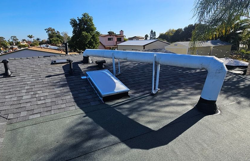 Rooftop view with dark shingles, vents, a skylight, and white pipes, with houses and blue sky in the background.