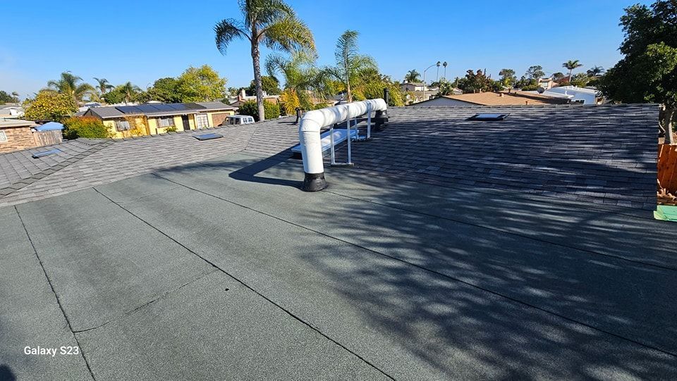Flat roof with vents and pipes, surrounded by suburban rooftops under a blue sky.