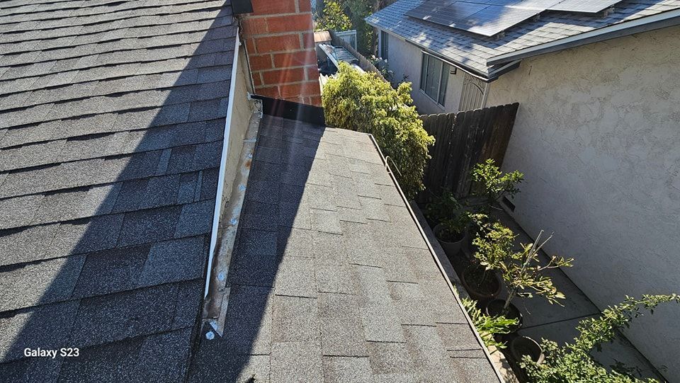 Overhead view of a roof with shingles, chimney, and a gutter filled with debris next to a fence.