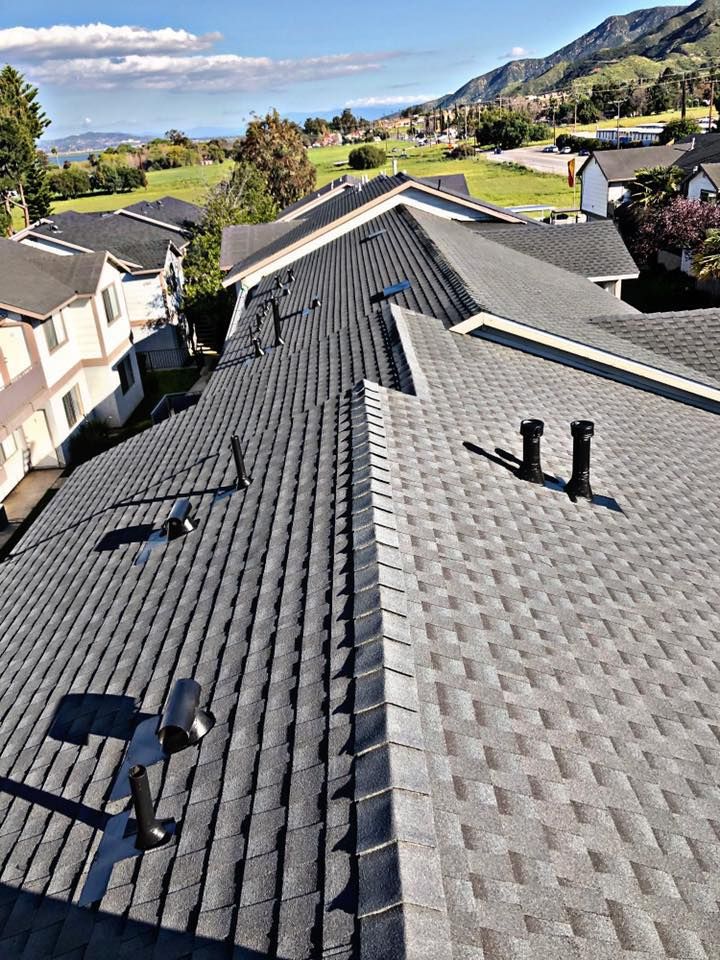 Overhead view of several dark gray shingled roofs with vents, in a suburban neighborhood with mountain backdrop.